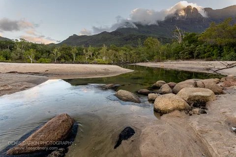 A still tidal channel cuts between sandbanks at Little Ramsey Bay, mirroring a cloud-wrapped mountain range in the last warm light of evening.