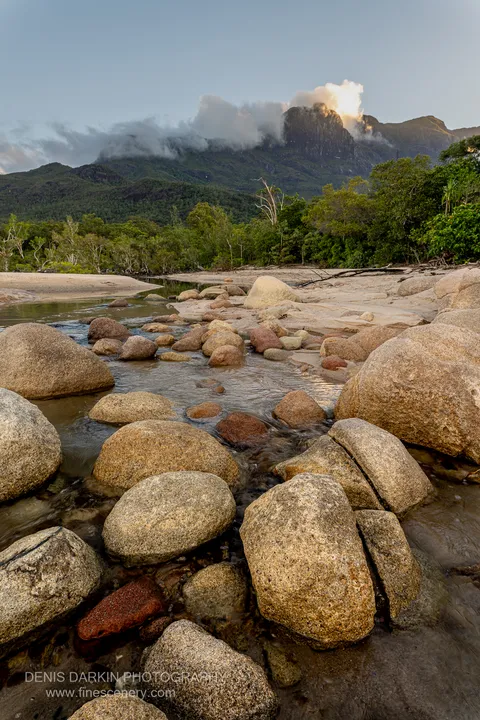 A clear stream winds between sun-warmed granite boulders at Little Ramsey Bay, with a craggy ridge and coastal forest rising behind in warm afternoon light.