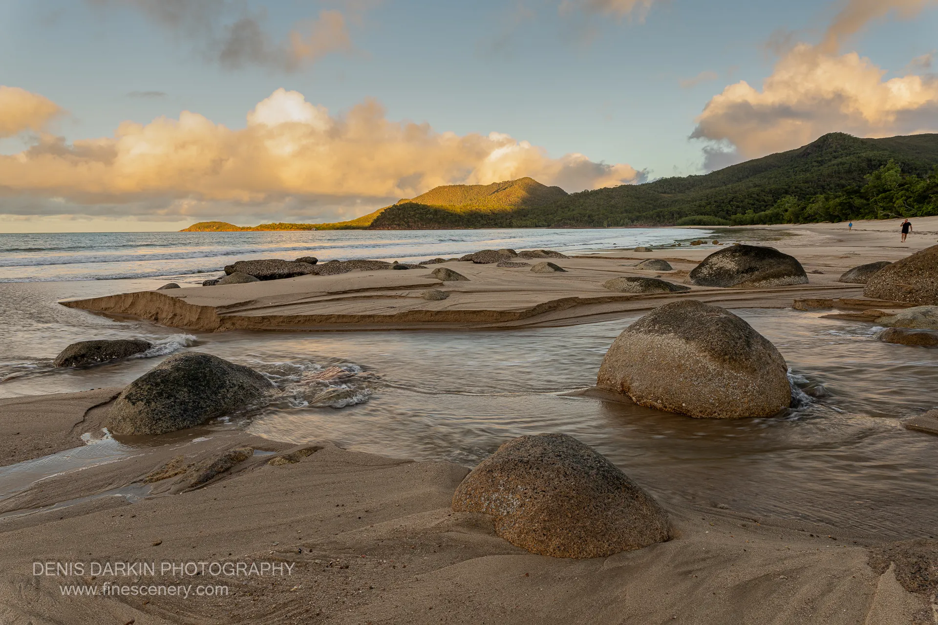 Low cloud sits above a forested headland at Little Ramsey Bay as wet sand and scattered granite boulders catch the last light along the shore.