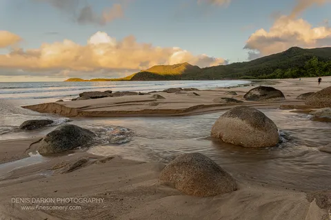 Low cloud sits above a forested headland at Little Ramsey Bay as wet sand and scattered granite boulders catch the last light along the shore.
