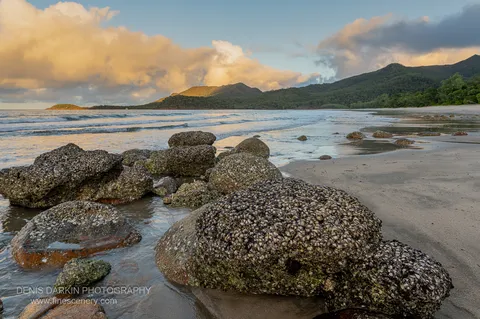 Barnacle- and oyster-crusted boulders at the tide line of Little Ramsey Bay with warm cloud catching last light above the headland.