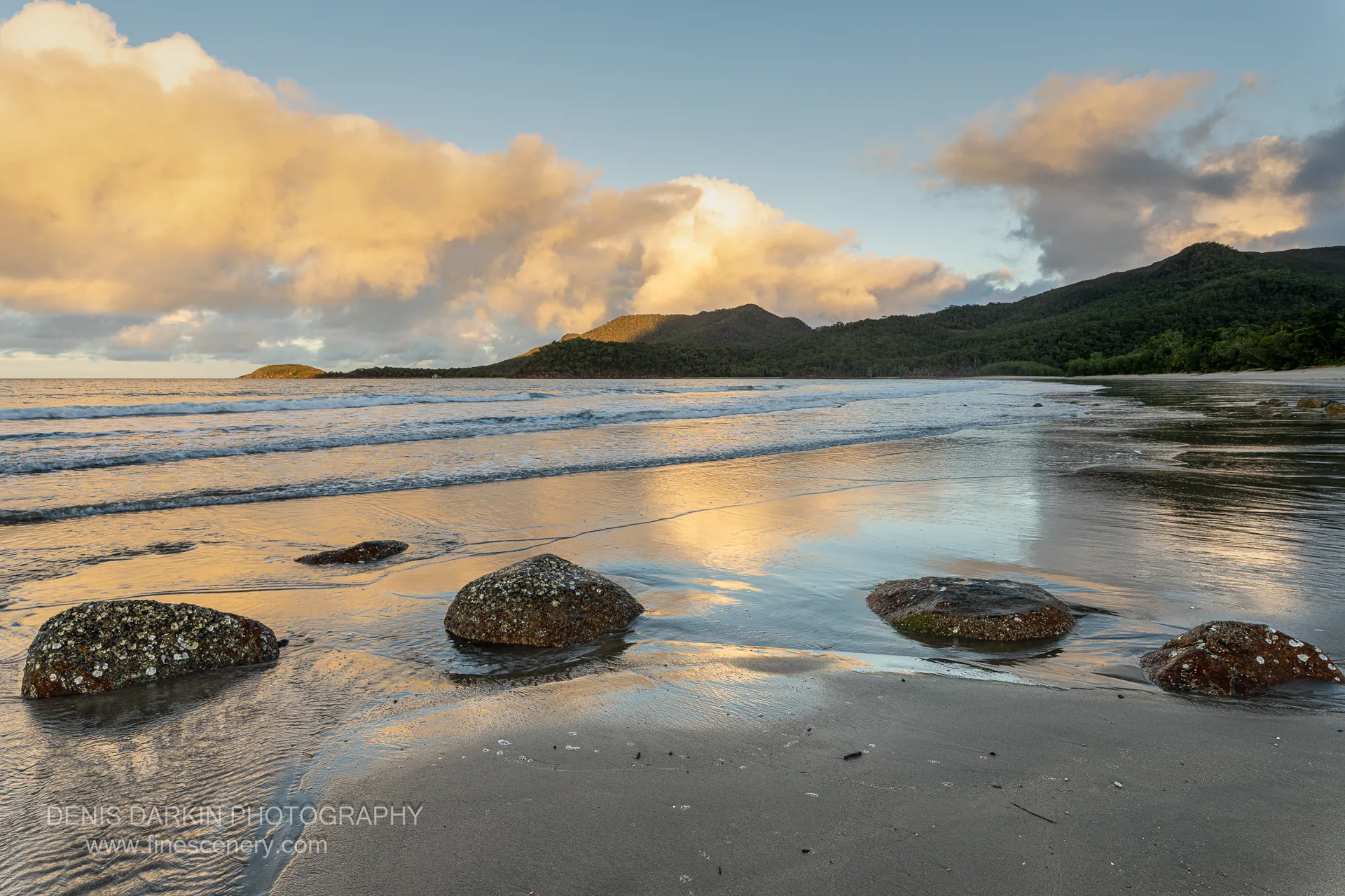 Four lichen-crusted boulders sit on the wet sand of Little Ramsey Bay as a retreating tide mirrors peach cloud overhead, forested headlands closing the bay.