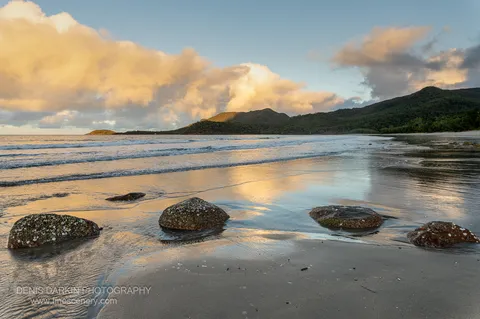 Four lichen-crusted boulders sit on the wet sand of Little Ramsey Bay as a retreating tide mirrors peach cloud overhead, forested headlands closing the bay.