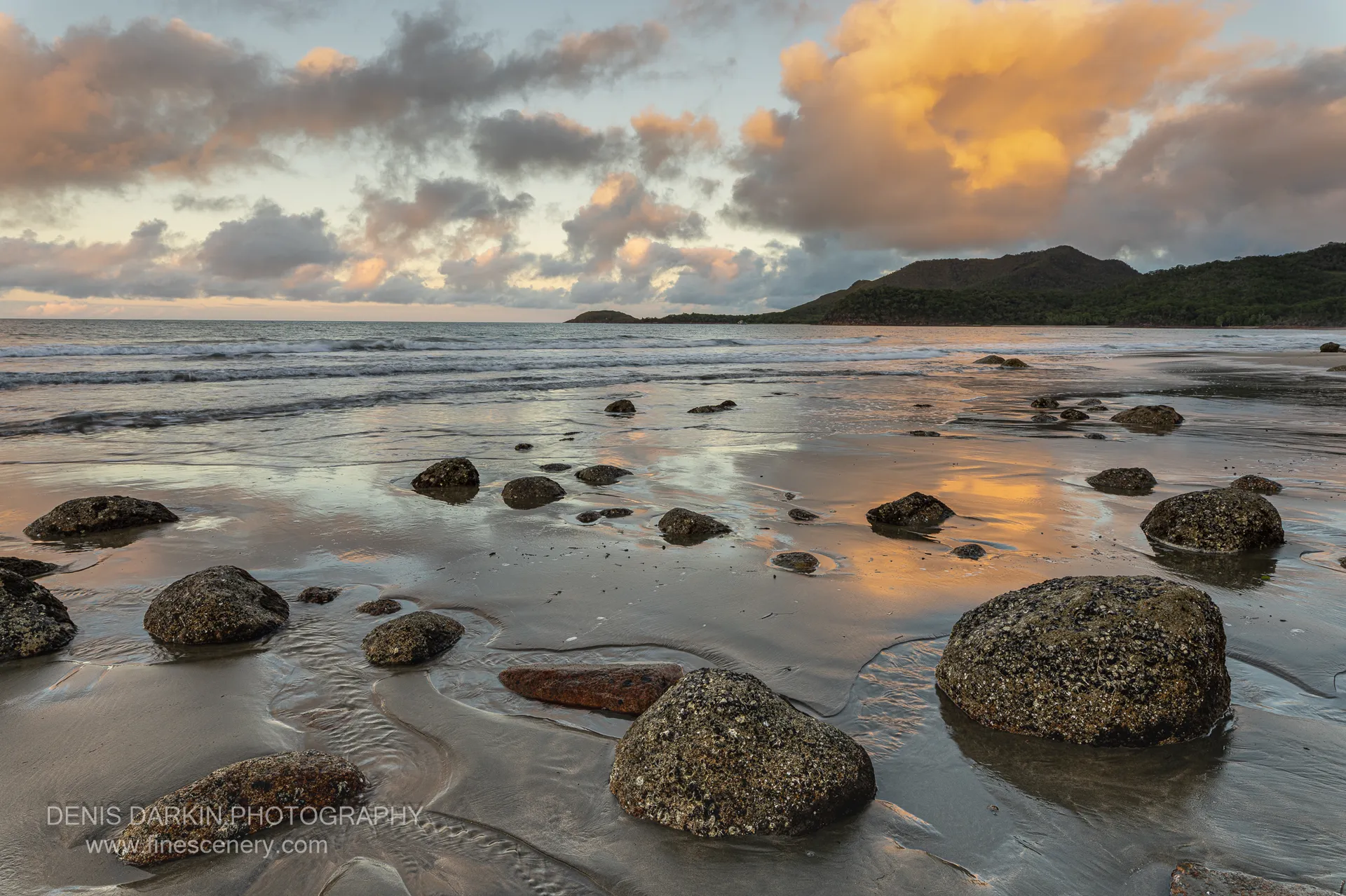 A shallow creek outflow threads around granite boulders at Little Ramsey Bay as two figures walk the far sand below a forested headland at sunset.