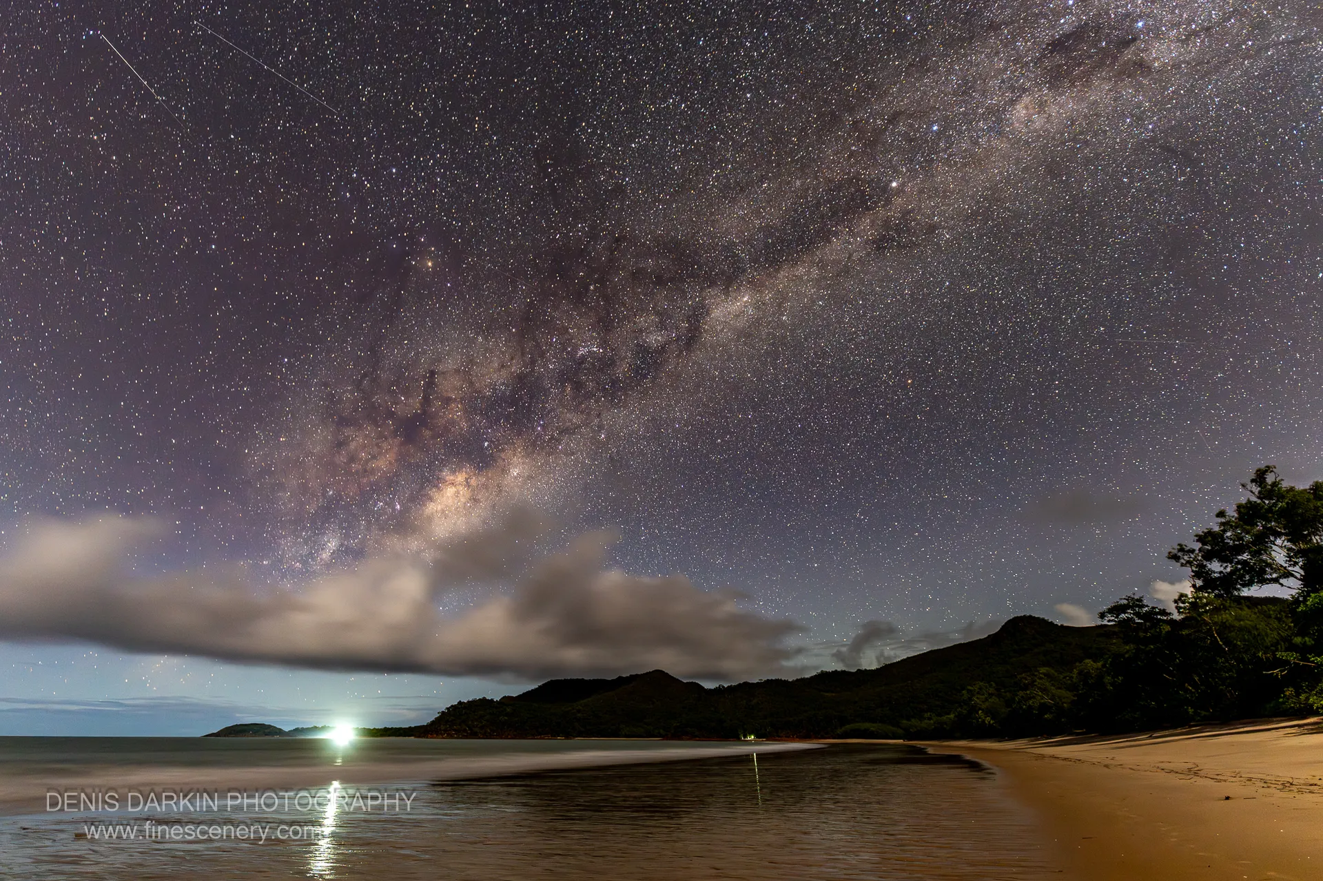 The Milky Way arcs over a dark headland at Little Ramsey Bay, its galactic core visible above low cloud and wet sand.