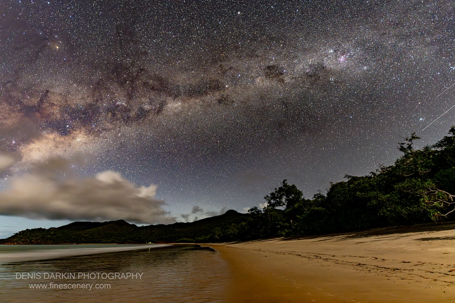 The Milky Way and galactic core arch over the dark headland and curved beach of Little Ramsey Bay on a winter night.