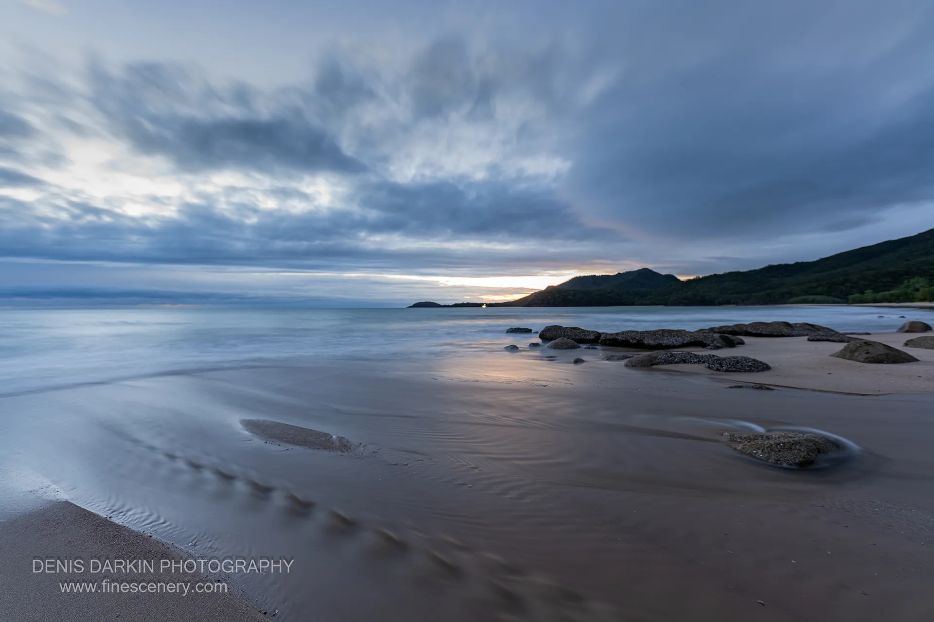 Long-exposure dawn at Little Ramsey Bay, with barnacle rocks on rippled sand and an orange band of light at the horizon behind a dark headland.