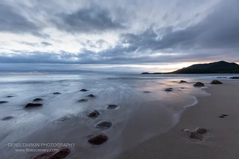 A line of dark boulders trails along wet sand at Little Ramsey Bay into long-exposure blue-grey surf beneath a streaked overcast sky.