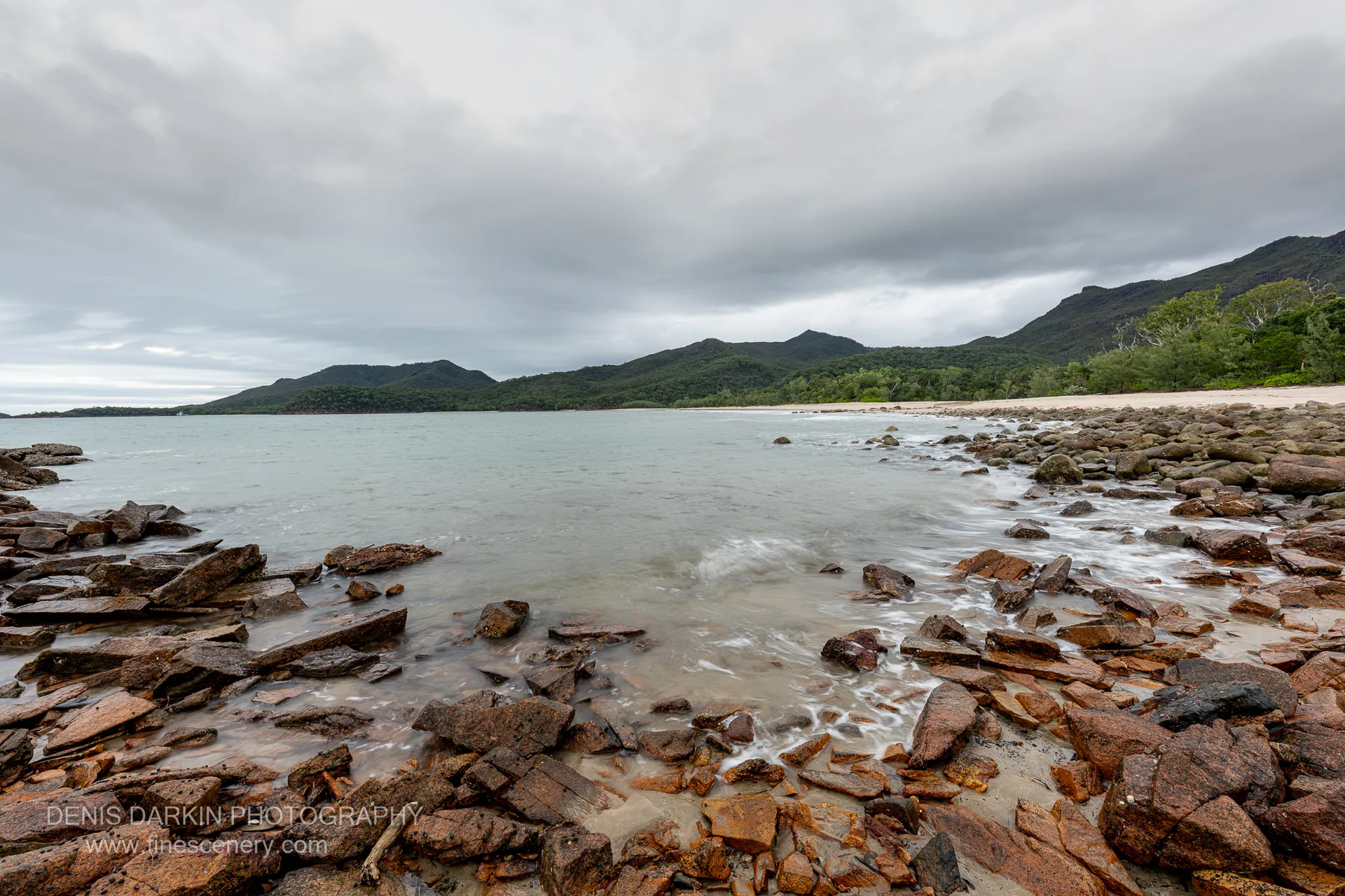 Rust-coloured rock shelf and angular boulders at Little Ramsey Bay, with long-exposure surf washing white toward forested ridgelines under an overcast sky.