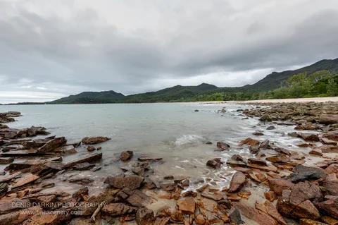 Rust-coloured rock shelf and angular boulders at Little Ramsey Bay, with long-exposure surf washing white toward forested ridgelines under an overcast sky.