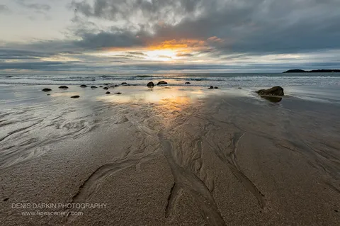 Branching wet-sand drainage channels at Little Ramsey Bay lead toward a muted sunrise, heavy cloud overhead with a thin orange gap near the horizon.
