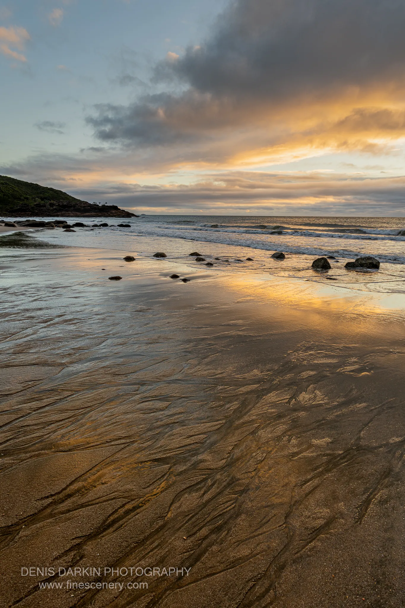 Fine drainage channels spread across golden wet sand at Little Ramsey Bay at sunrise, leading toward scattered rocks and a streaked orange sky.