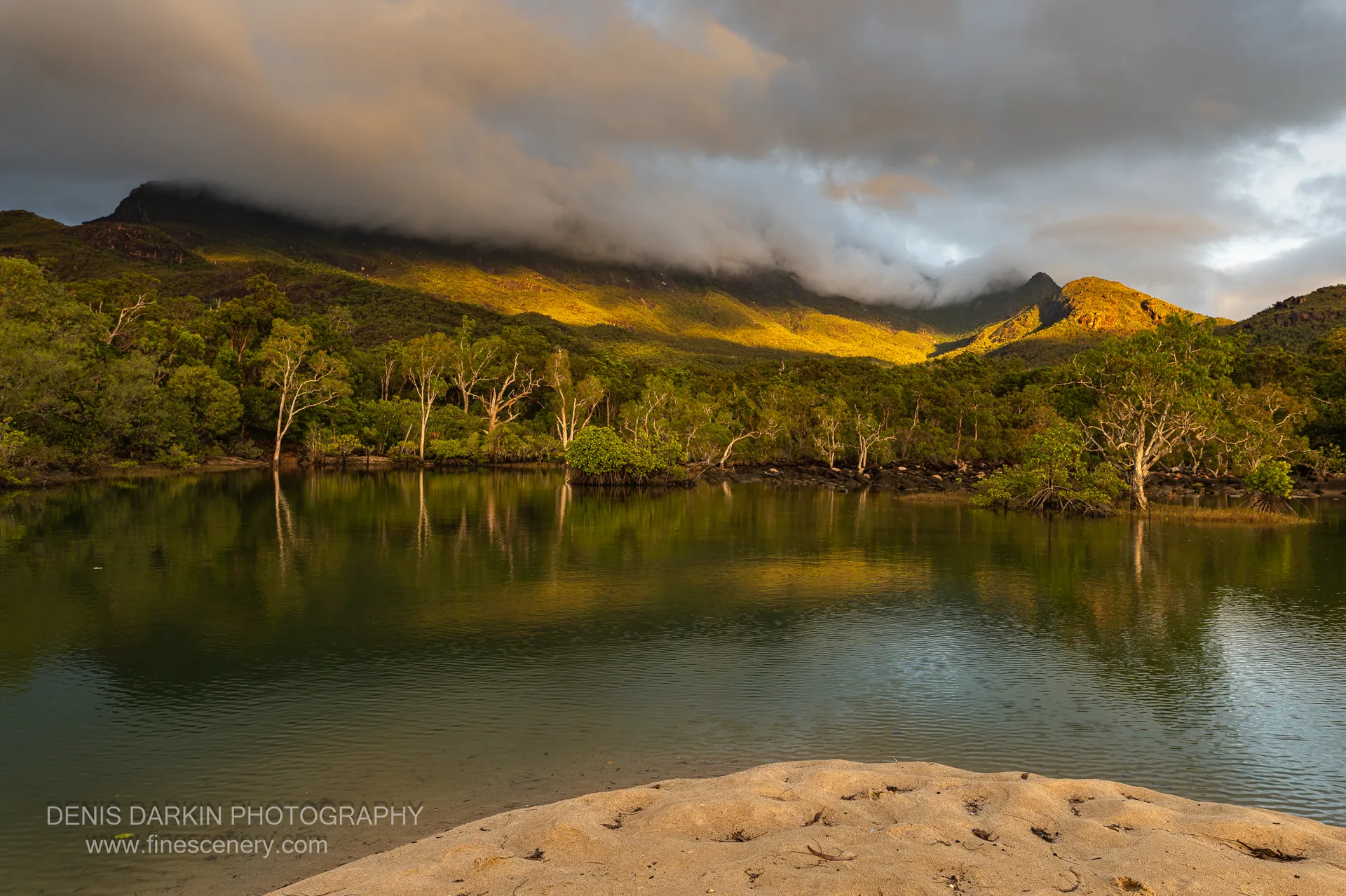 A still lagoon at Little Ramsey Bay reflects a mangrove-fringed forested ridge under a heavy cloud bank in early morning light.