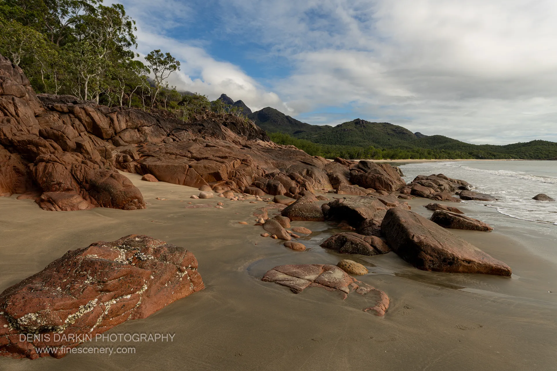 Weathered red boulders streaked with lichen crowd a beach corner on Hinchinbrook Island, with a forested range and partial blue sky beyond.