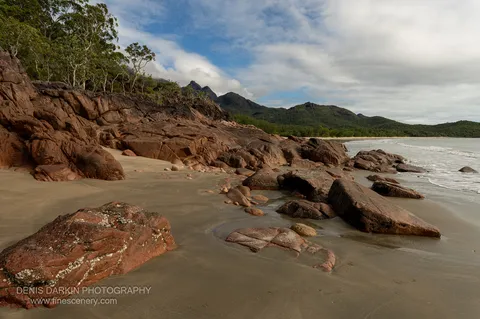 Weathered red boulders streaked with lichen crowd a beach corner on Hinchinbrook Island, with a forested range and partial blue sky beyond.