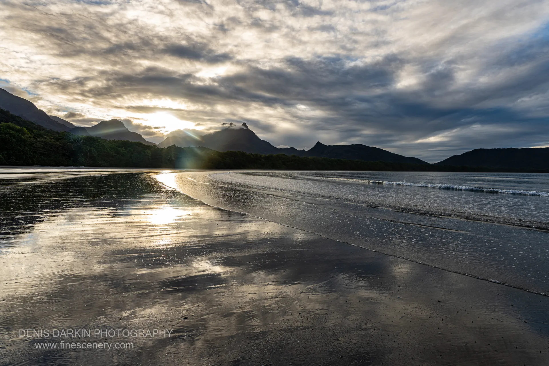 Low sun breaks through cloud behind the silhouetted Hinchinbrook range at Zoe Bay, with a starburst reflection running down wet sand at golden hour.