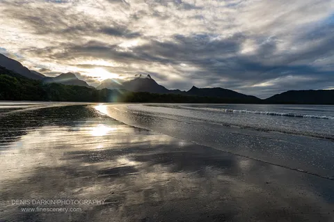 Low sun breaks through cloud behind the silhouetted Hinchinbrook range at Zoe Bay, with a starburst reflection running down wet sand at golden hour.