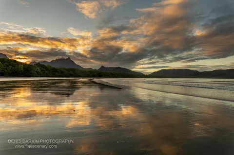 Receding water on the wide tidal flat of Zoe Bay mirrors orange-lit cloud at sunset, with a cloud-shrouded peak beyond.