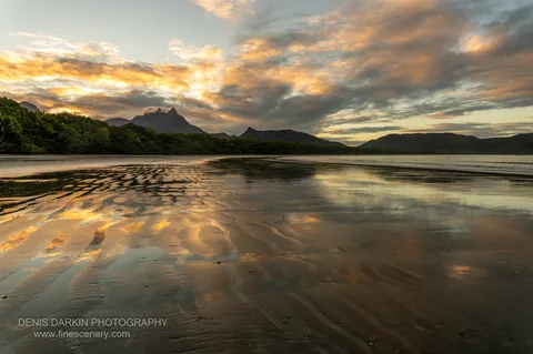 Ridged wet sand at low tide on Zoe Bay reflects orange-lit cloud and a cloud-topped peak, with dark forested headlands beyond.