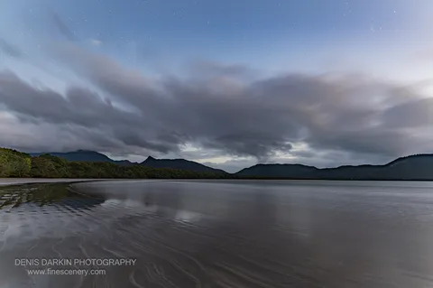 Before dawn at Zoe Bay, a long exposure smooths the tidal flat and bay beneath a heavy cloud bank drifting over low ranges.