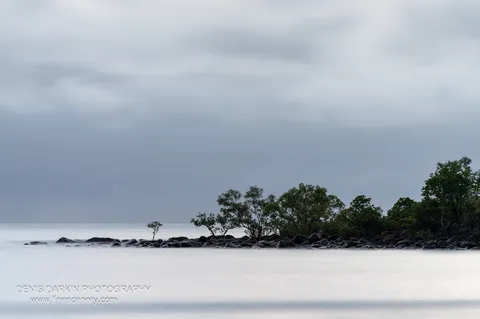 A rocky spit topped with wind-bent mangroves and shrubs sits between long-exposure sea and an overcast sky at dawn near Zoe Bay.