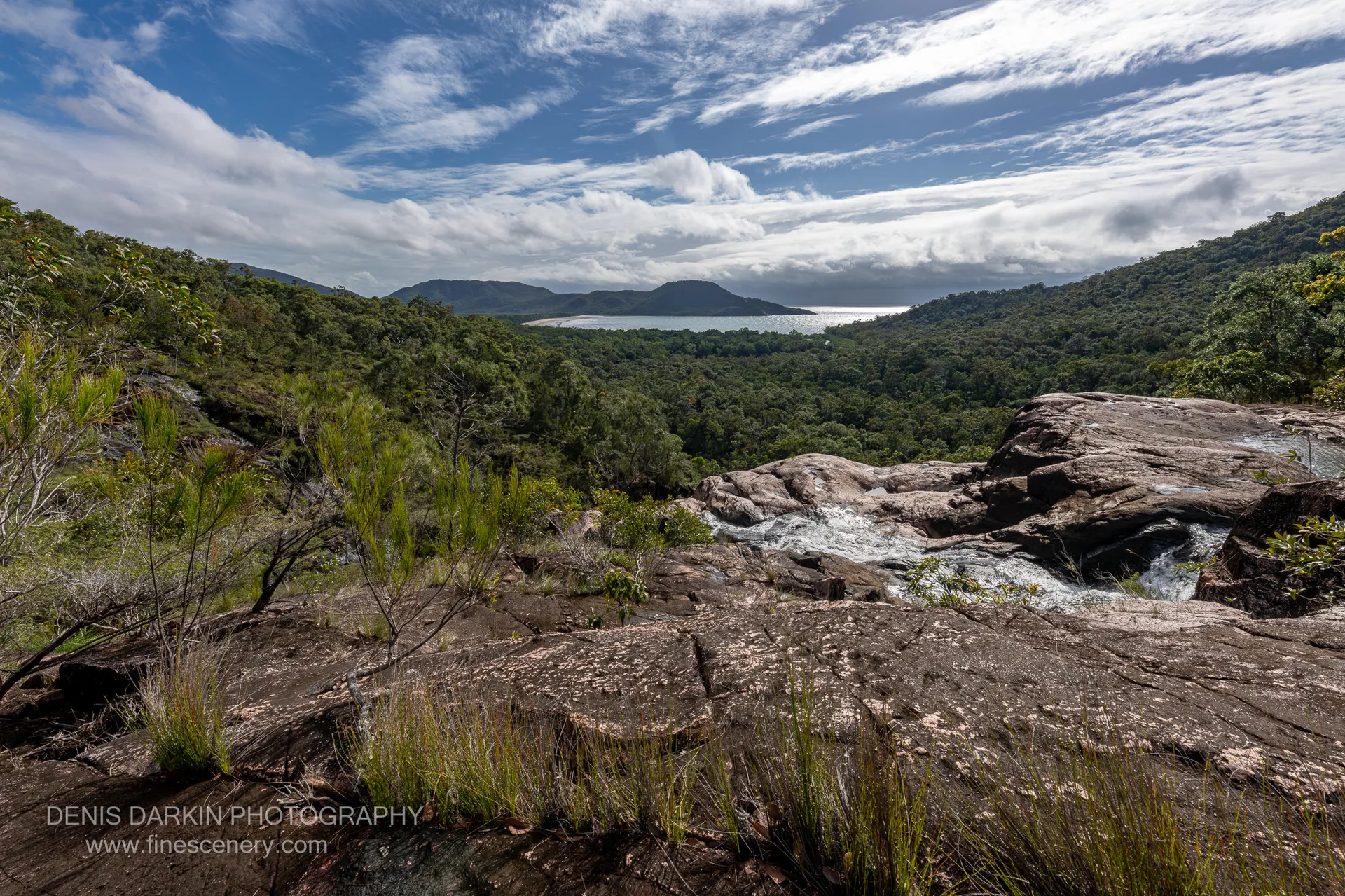 Water slides over pink granite slabs at the top of Zoe Falls, with Zoe Bay's beach and the sea visible through the forested valley below.