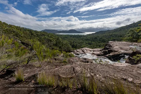 Water slides over pink granite slabs at the top of Zoe Falls, with Zoe Bay's beach and the sea visible through the forested valley below.
