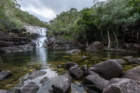 A low waterfall slides down a smooth granite slab into a clear plunge pool ringed by dark boulders and rainforest at Mulligan Falls, Hinchinbrook Island.