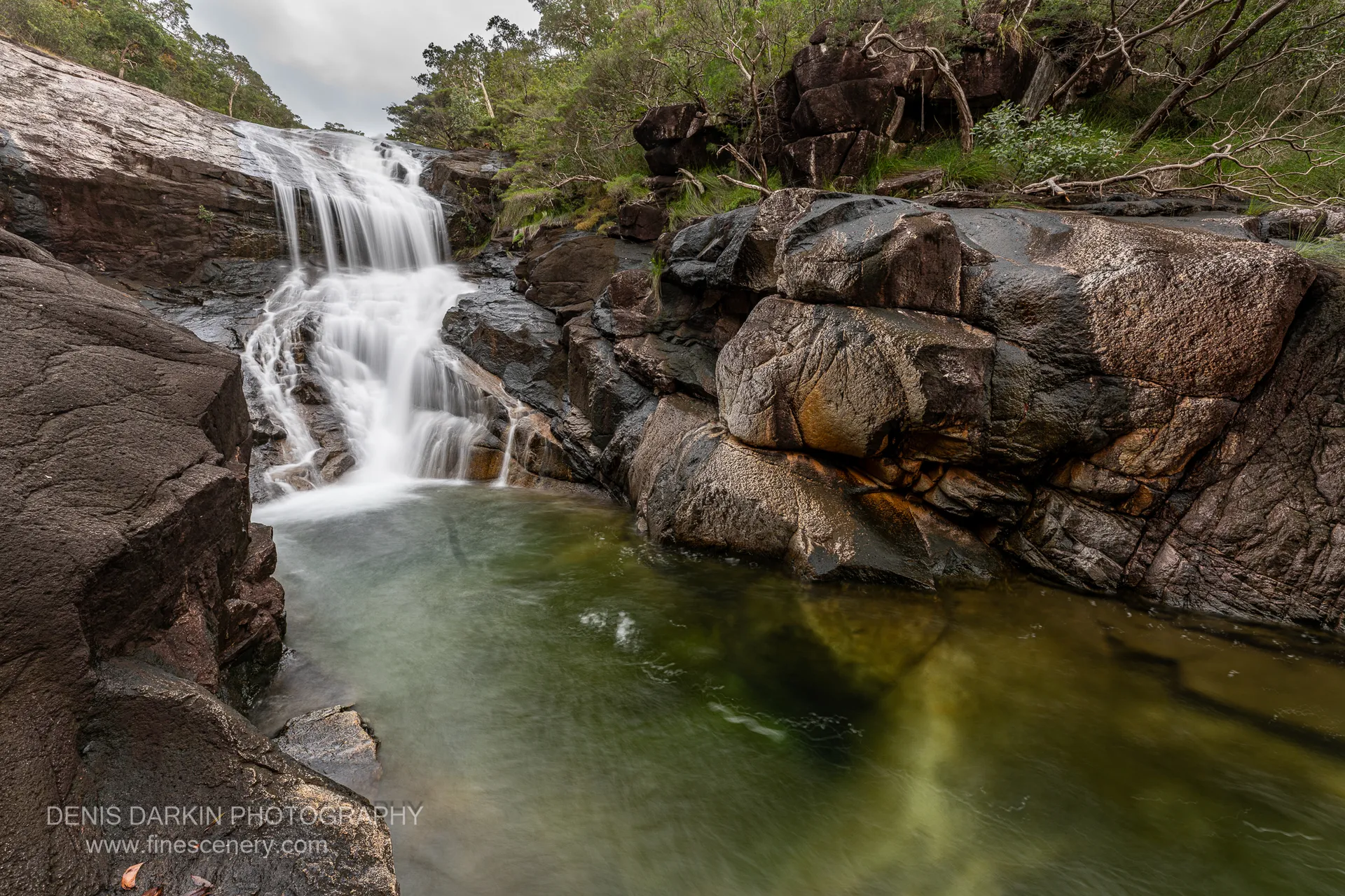 Mulligan Falls fans across a tiered rock slab into a green plunge pool surrounded by large rounded boulders and rainforest, under overcast light.