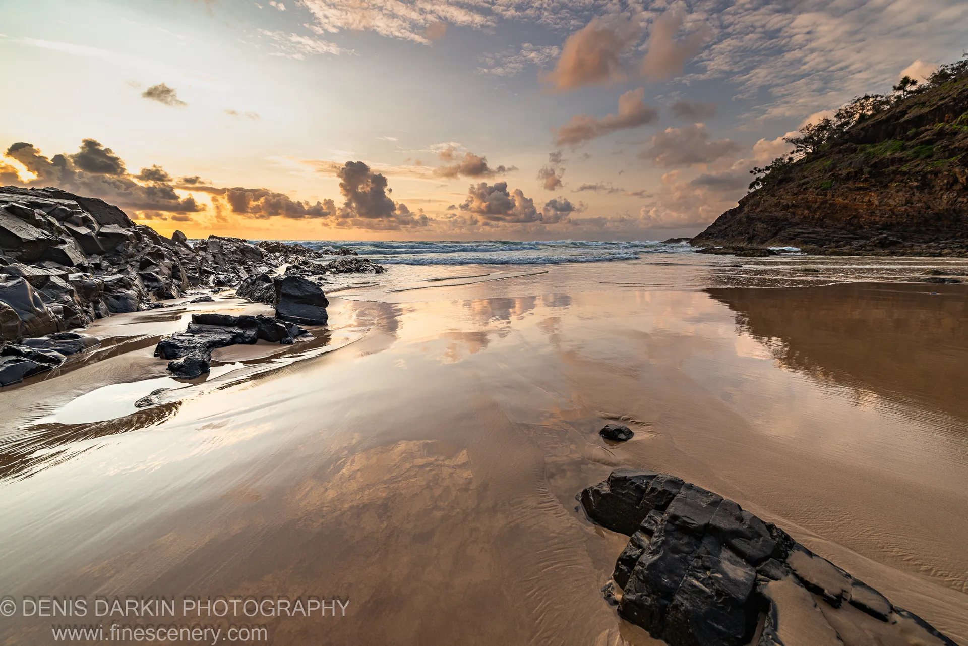 Warm dawn light reflects in pools on wet sand at low tide, with dark angular rocks lining the Byron Bay shore.