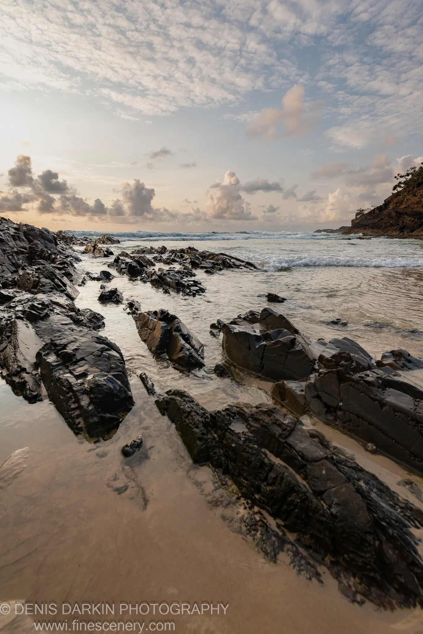 Glossy dark rocks form a diagonal line across a Queensland beach at dawn, leading toward breaking waves on the horizon under softly lit clouds.
