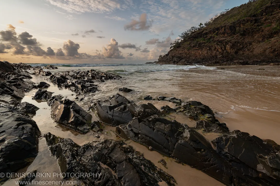 Dark volcanic rocks and shallow tide pools on a Queensland beach at dawn, with gentle waves and a sandy cove below a steep cliff.