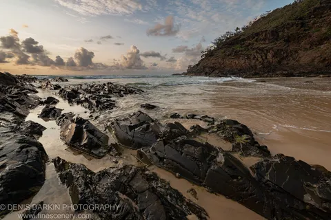 Dark volcanic rocks and shallow tide pools on a Queensland beach at dawn, with gentle waves and a sandy cove below a steep cliff.