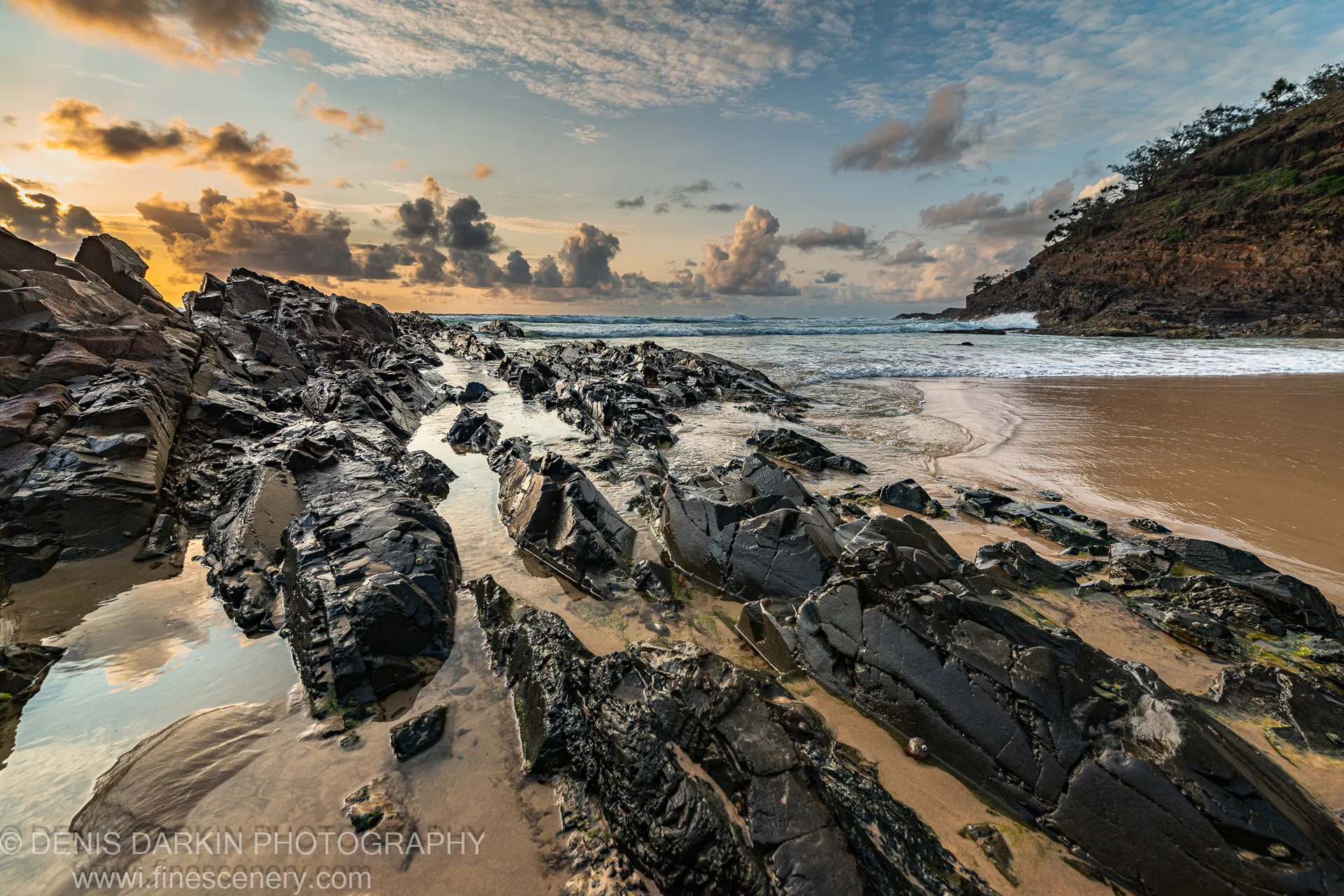 Dark glistening rock formations channel a trickle of seawater toward the sandy beach as sunrise light catches the tide pools.