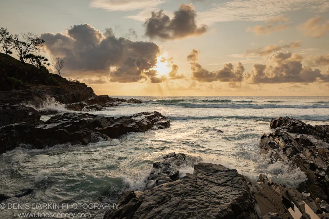 Dark rocks with white surf at their base on the Queensland coast, the sun breaking through cloud to cast a golden path across the sea.
