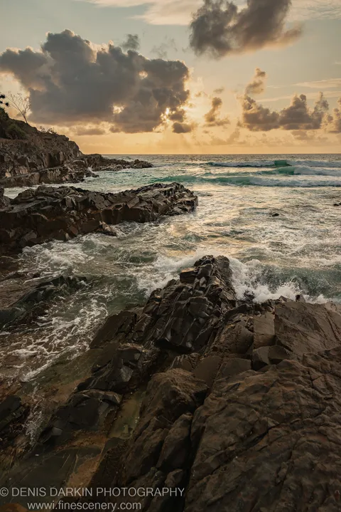 Jagged columnar rock formations wet from breaking waves, with morning light filtering through low cloud on the Queensland coast.