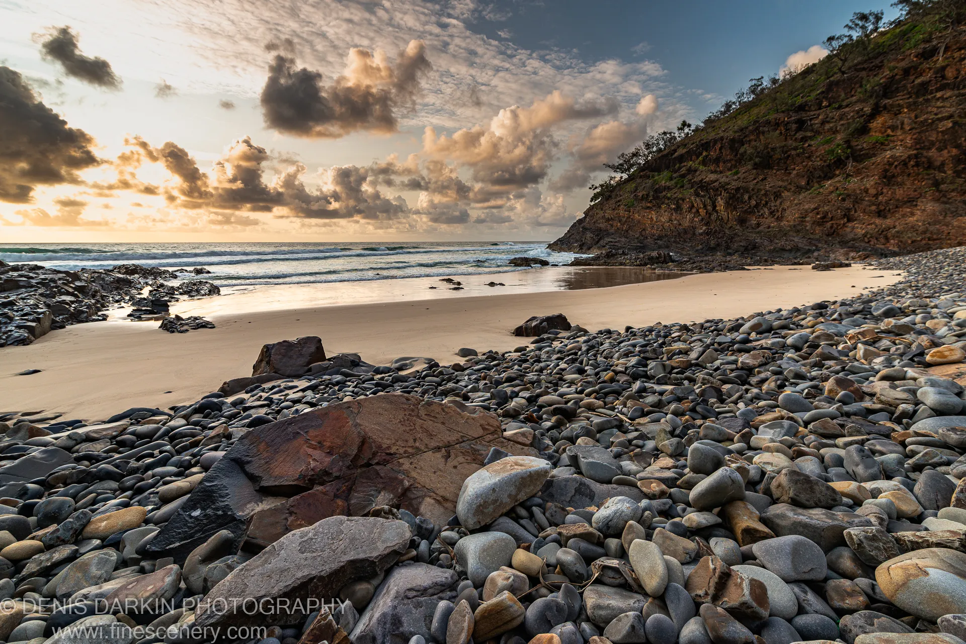 Sandy cove at dawn on the Queensland coast, with warm light across wet sand and a steep vegetated cliff to one side.