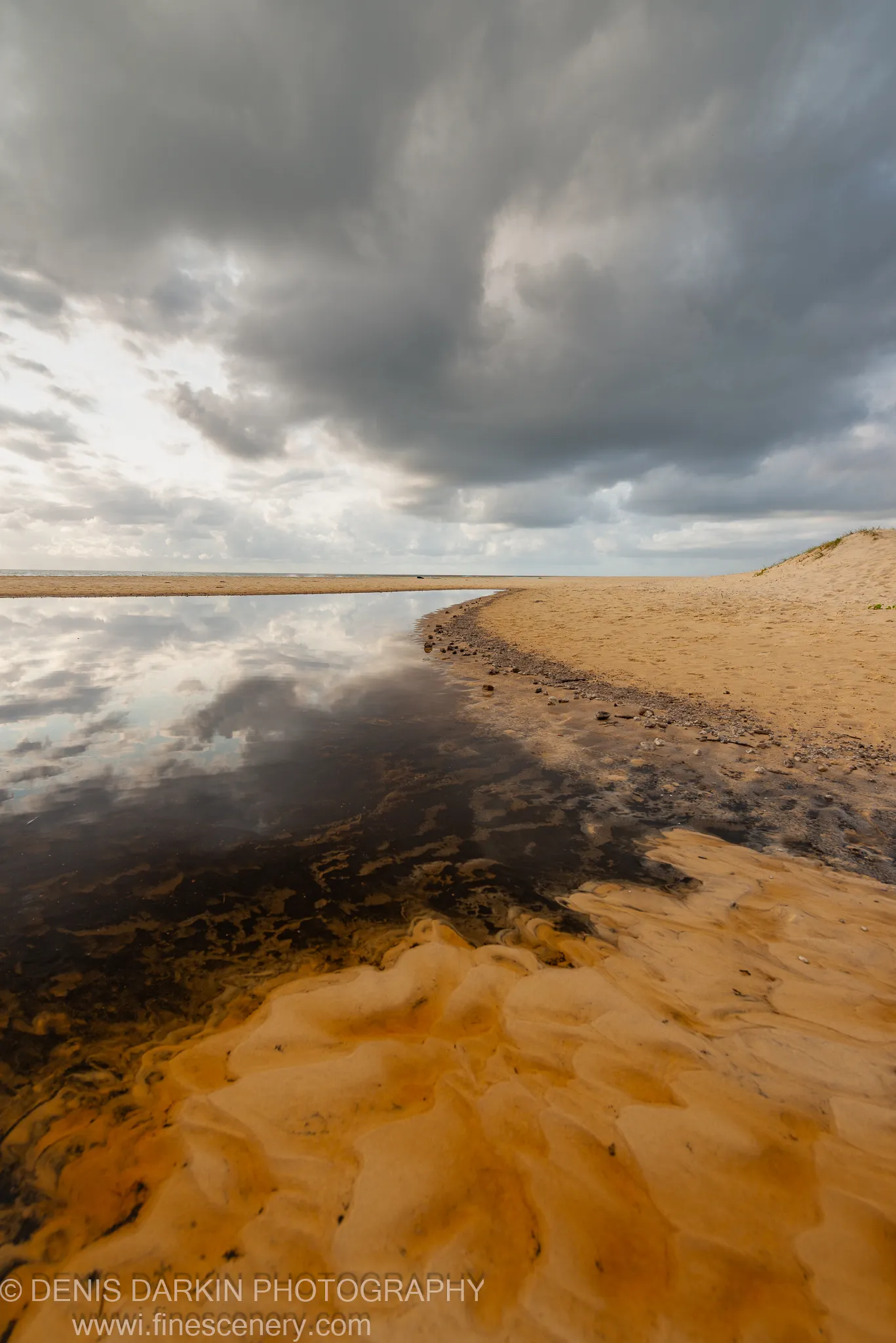 Wet sand textures in the foreground lead to a curved shoreline and tide pool reflecting a heavy sky on the Queensland coast.