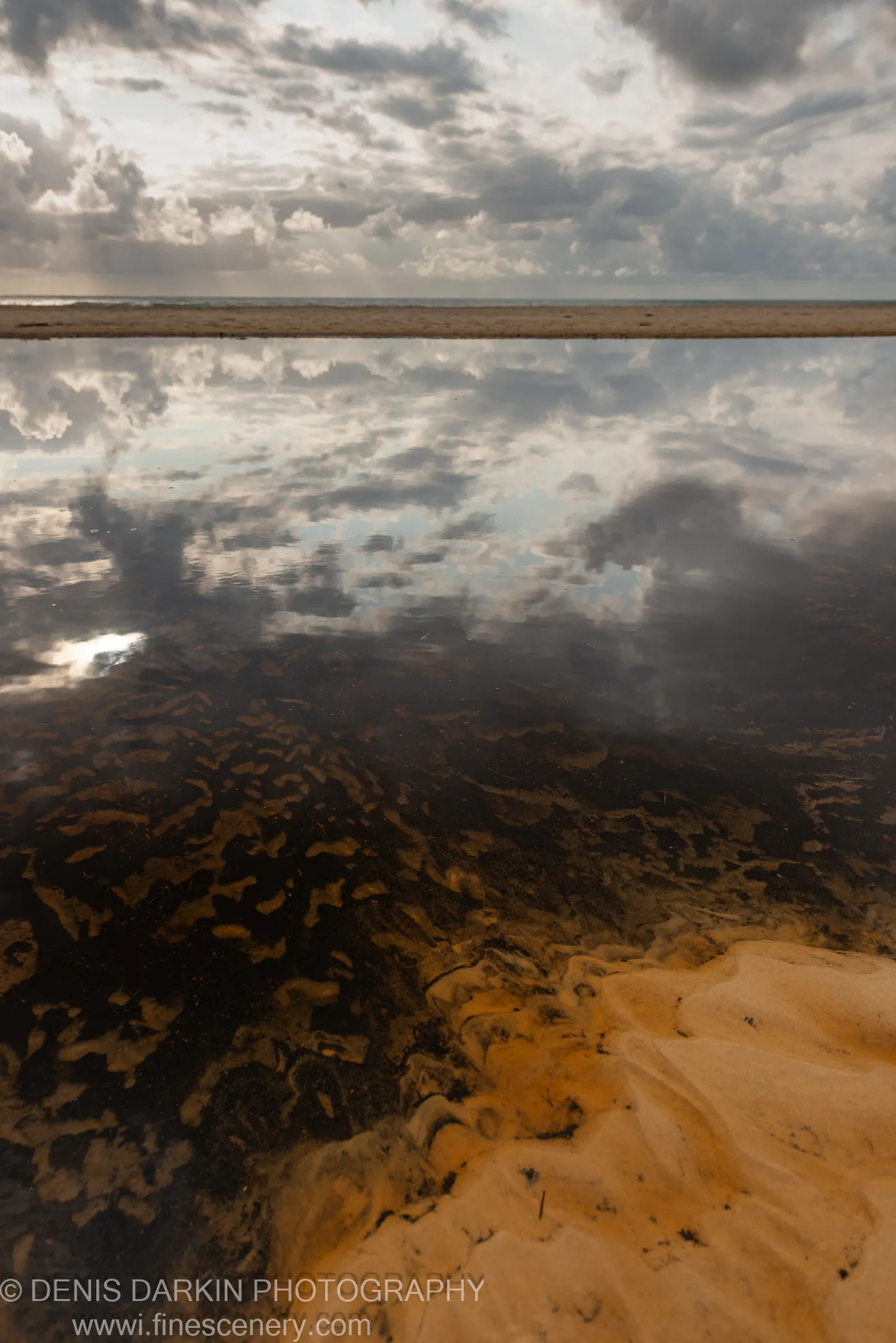 Rippled sand and dark algae beneath shallow water on a tidal flat, with sunbeams over the distant shore on the Queensland coast.