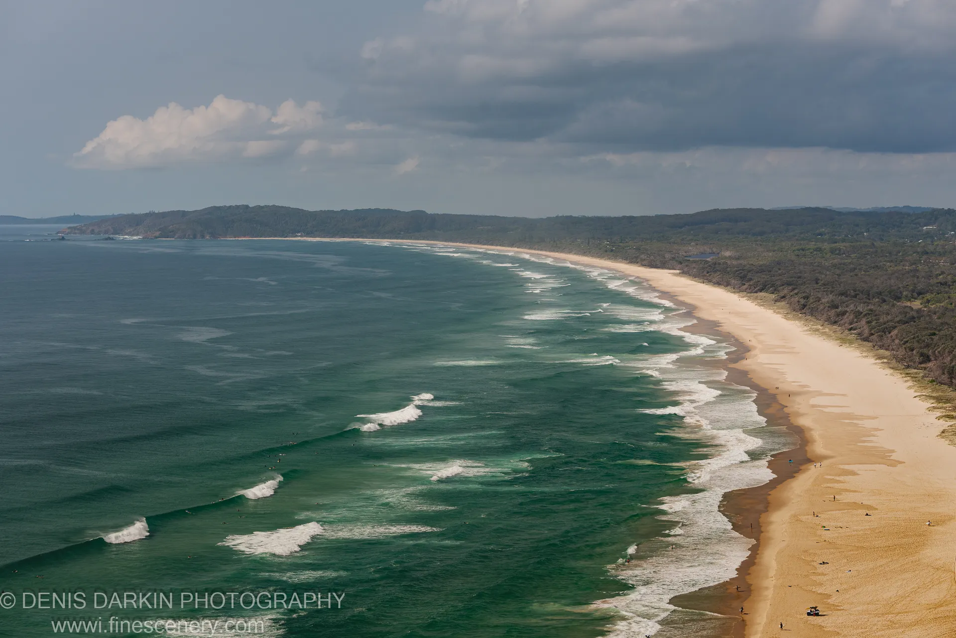 A crescent-shaped beach viewed from above on the Queensland coast, white surf breaking along the shoreline with tree-covered headlands in the distance.