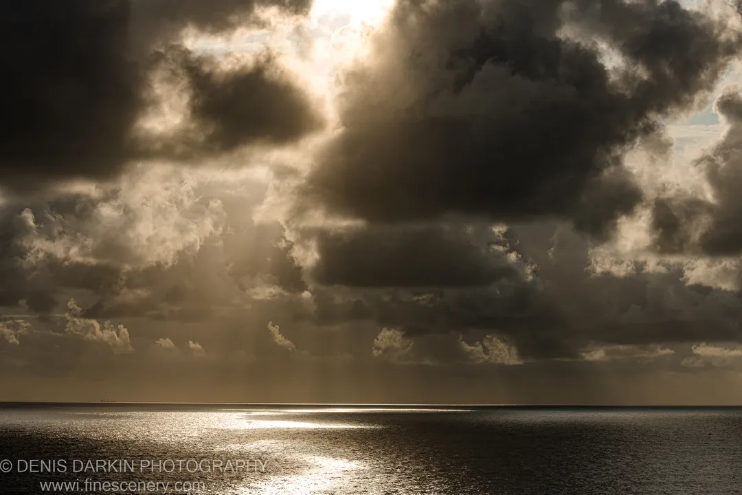 Shafts of dawn light break through heavy storm clouds over a dark, calm ocean off the Queensland coast.