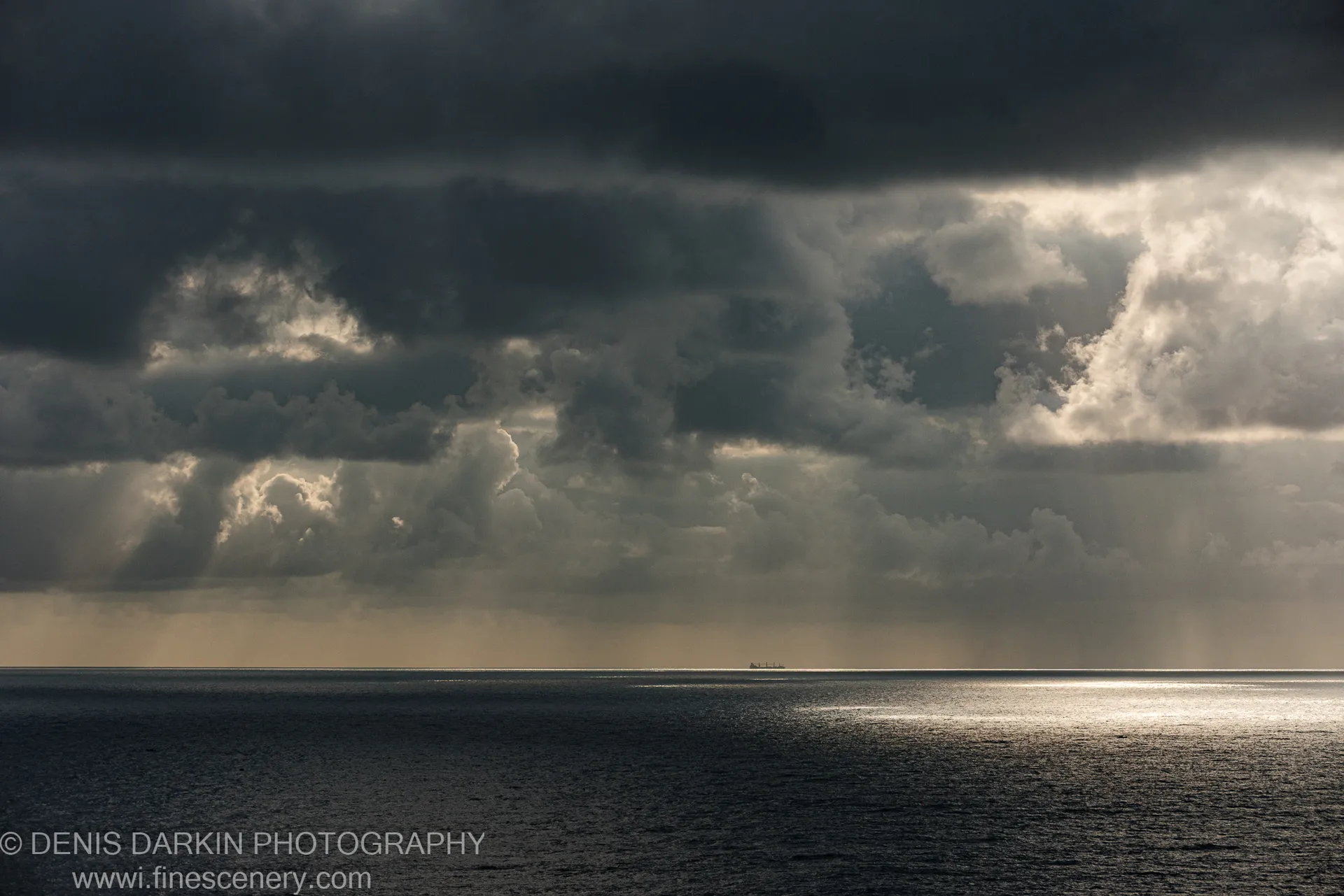 Dark storm clouds over a vast expanse of ocean at dawn, with rain curtains falling and a small ship silhouette on the horizon.