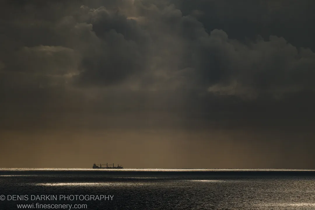 A cargo ship sits in telephoto silhouette against a textured wall of cumulus clouds over the Queensland coast at dawn.