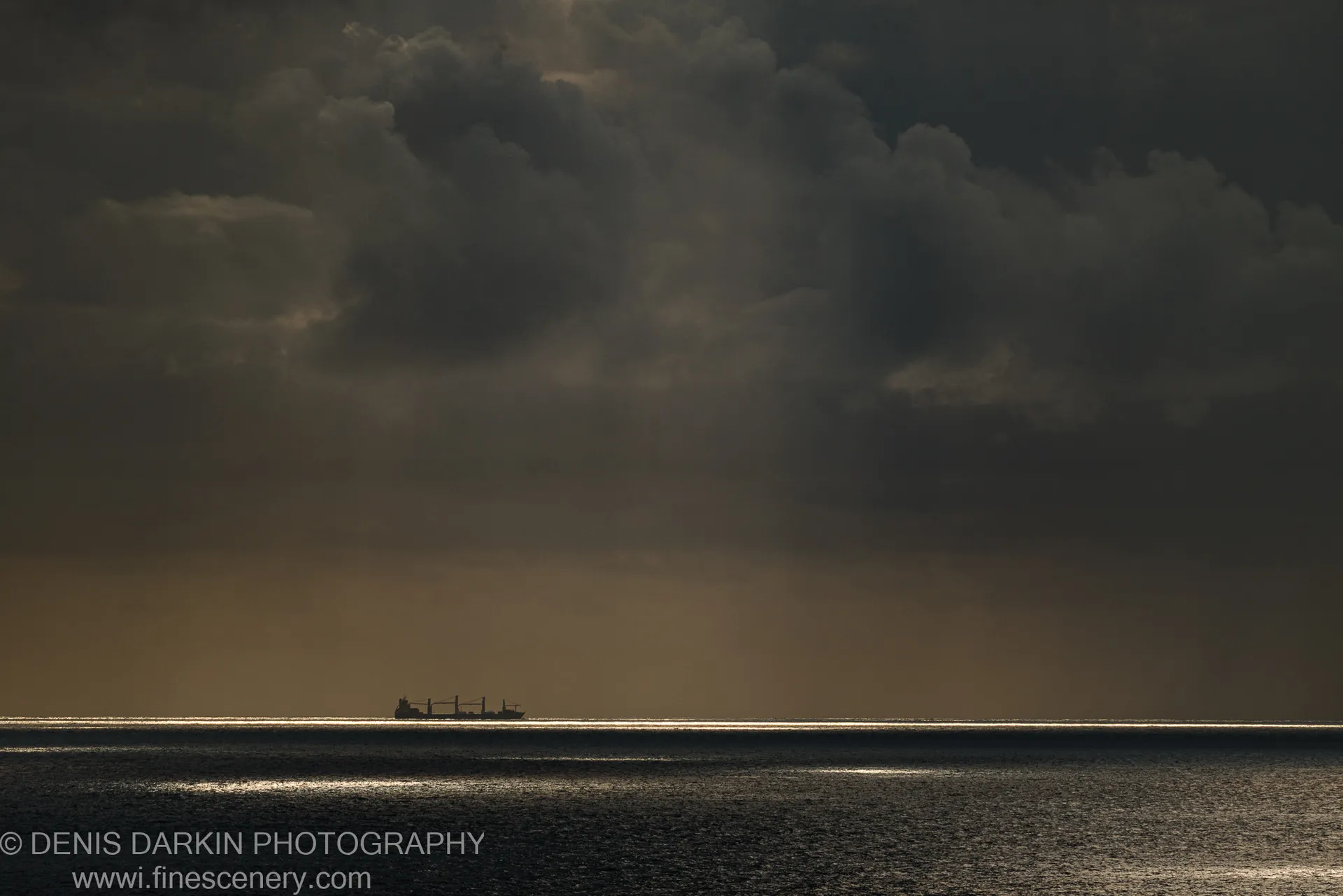 A cargo ship sits in telephoto silhouette against a textured wall of cumulus clouds over the Queensland coast at dawn.