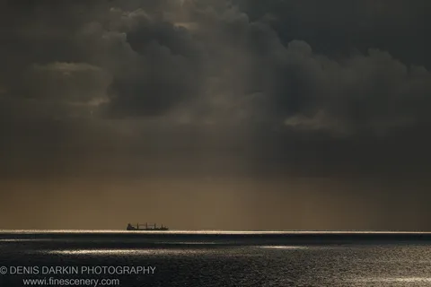 A cargo ship sits in telephoto silhouette against a textured wall of cumulus clouds over the Queensland coast at dawn.
