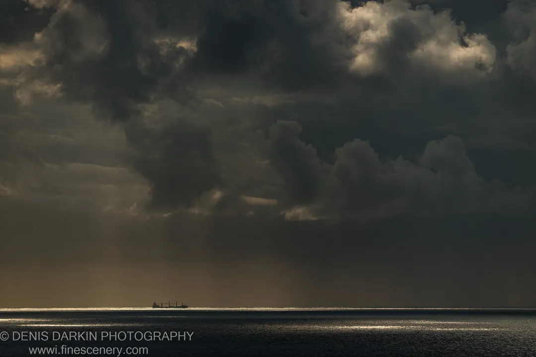 A distant cargo vessel is silhouetted against a narrow band of warm horizon light beneath heavy cumulus clouds over the ocean.