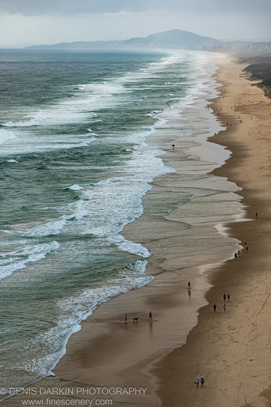 Breaking surf lines cut diagonally across a Queensland beach viewed from above, with wet sand reflections and a hazy headland in the distance.