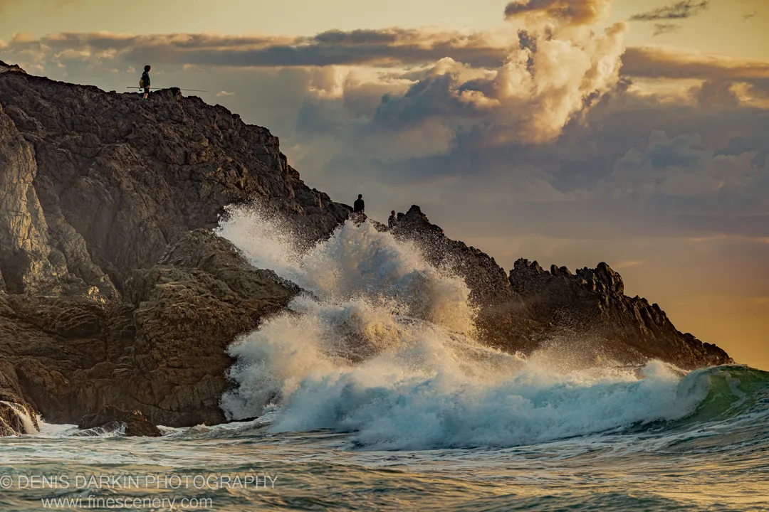A wave crashes against dark basalt cliffs at Coffs Harbor, sending a plume of white spray into the predawn sky.
