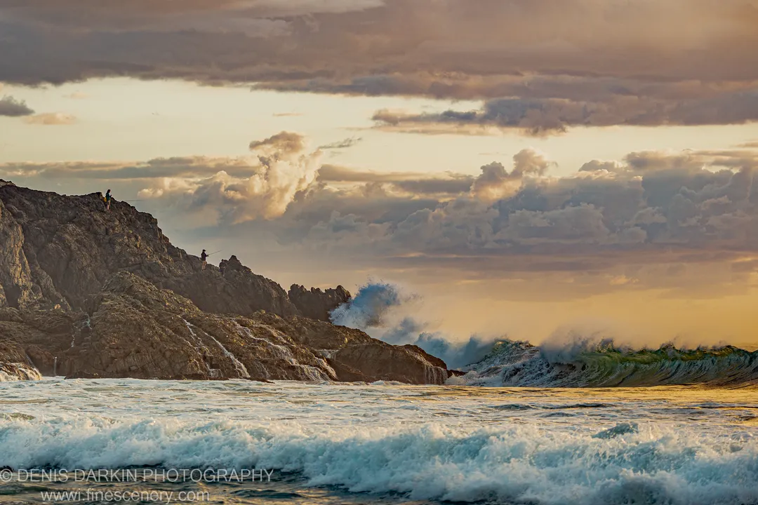 Three silhouetted figures stand on a high cliff at Coffs Harbor above crashing waves, with warm-lit clouds at the predawn horizon.