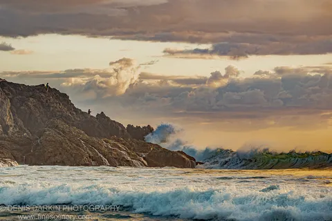 Three silhouetted figures stand on a high cliff at Coffs Harbor above crashing waves, with warm-lit clouds at the predawn horizon.